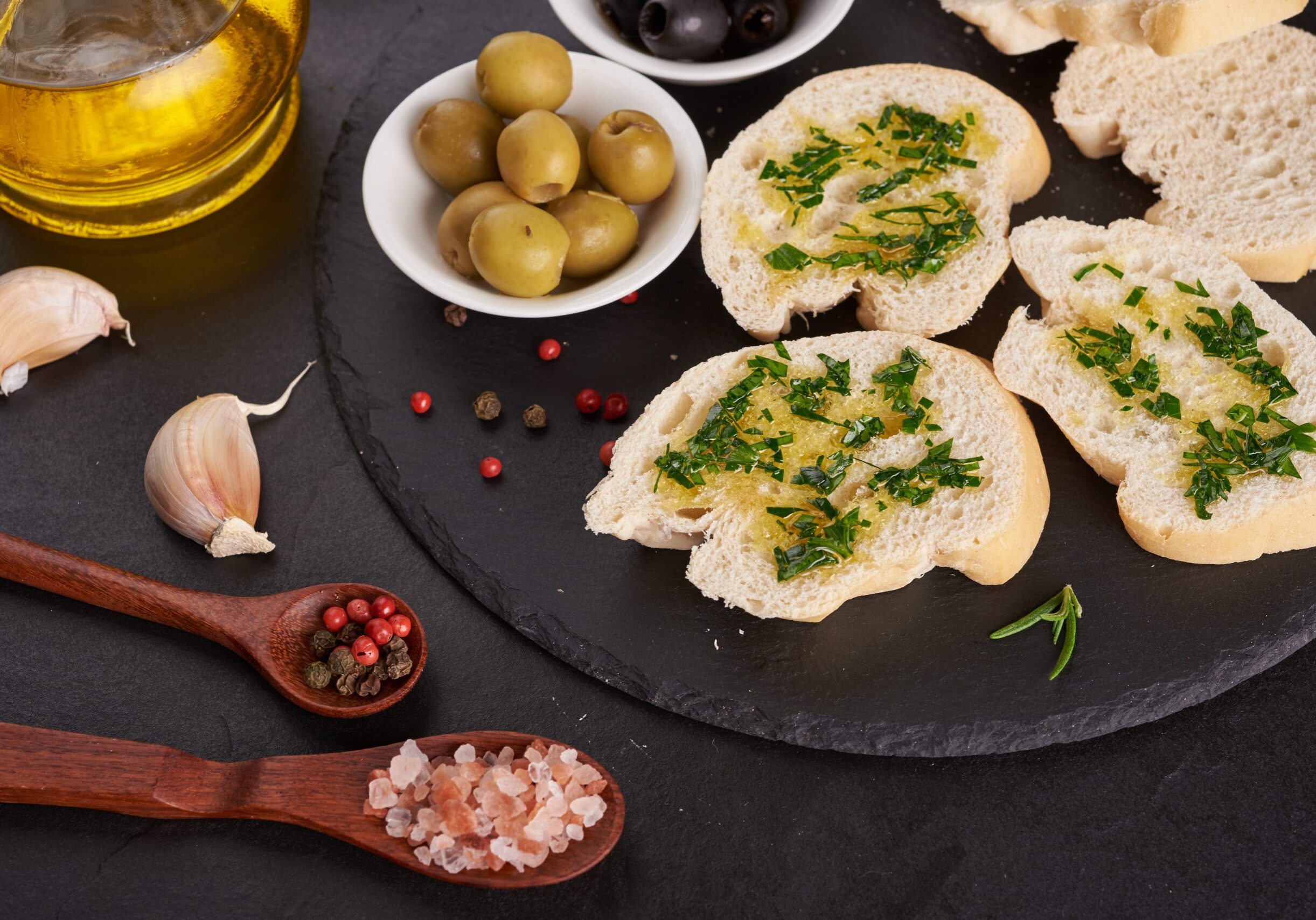 Mediterranean snacks set. Olives, oil, herbs and sliced ciabatta bread on black slate stone board over painted dark blue background, top view. Flat lay.